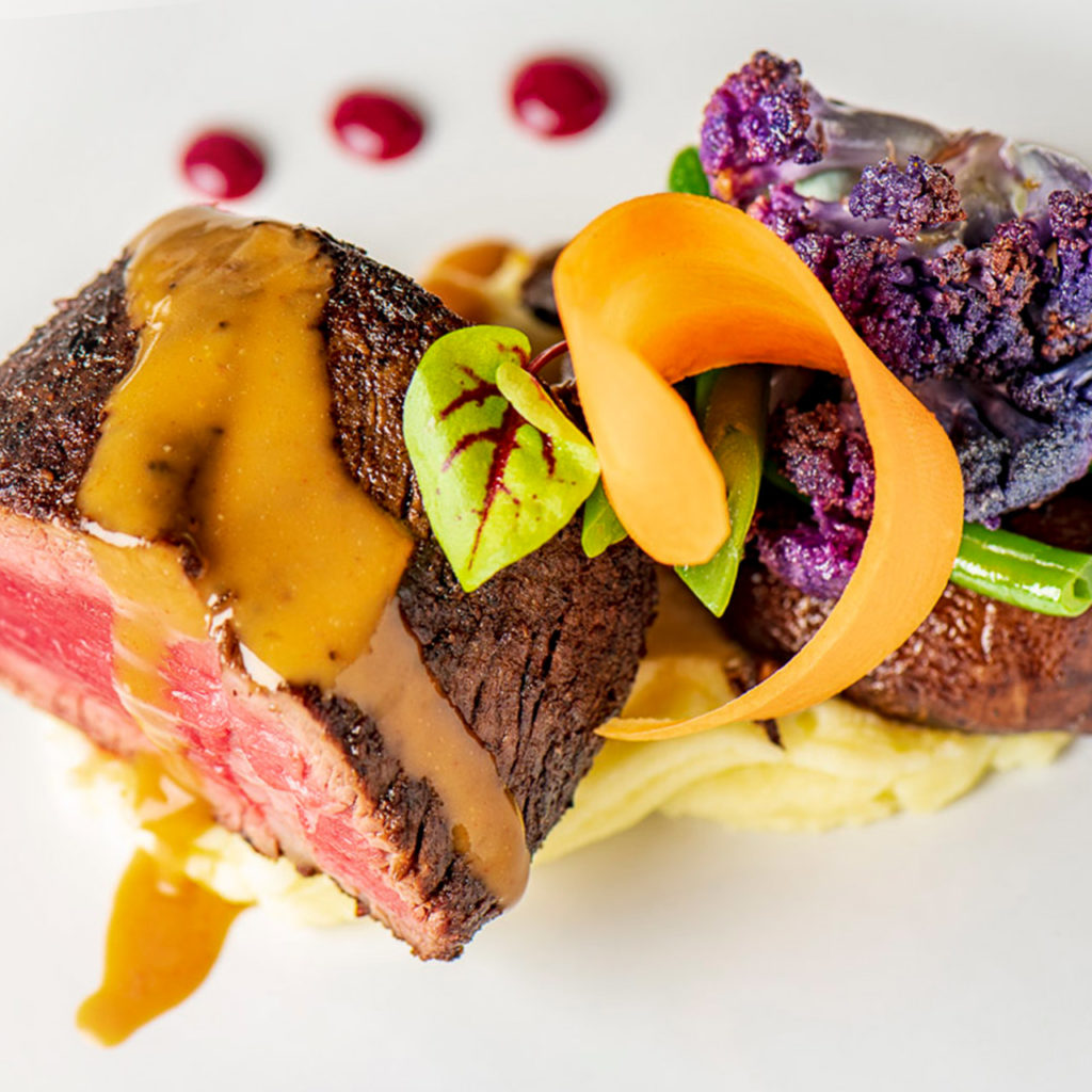 Sliced beef with sauce on mashed potatoes, garnished with carrot, greens, and purple cauliflower, with sauce dots on the side.