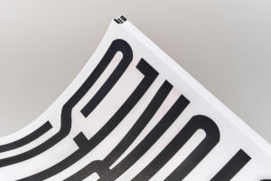 Close-up of a glossy booklet corner, showing oversized black text on a white page against a gray background.