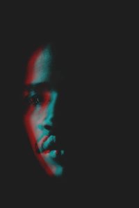 Portrait of a person in low light, partially illuminated with blue and red lighting, creating a dramatic contrast against the dark background.