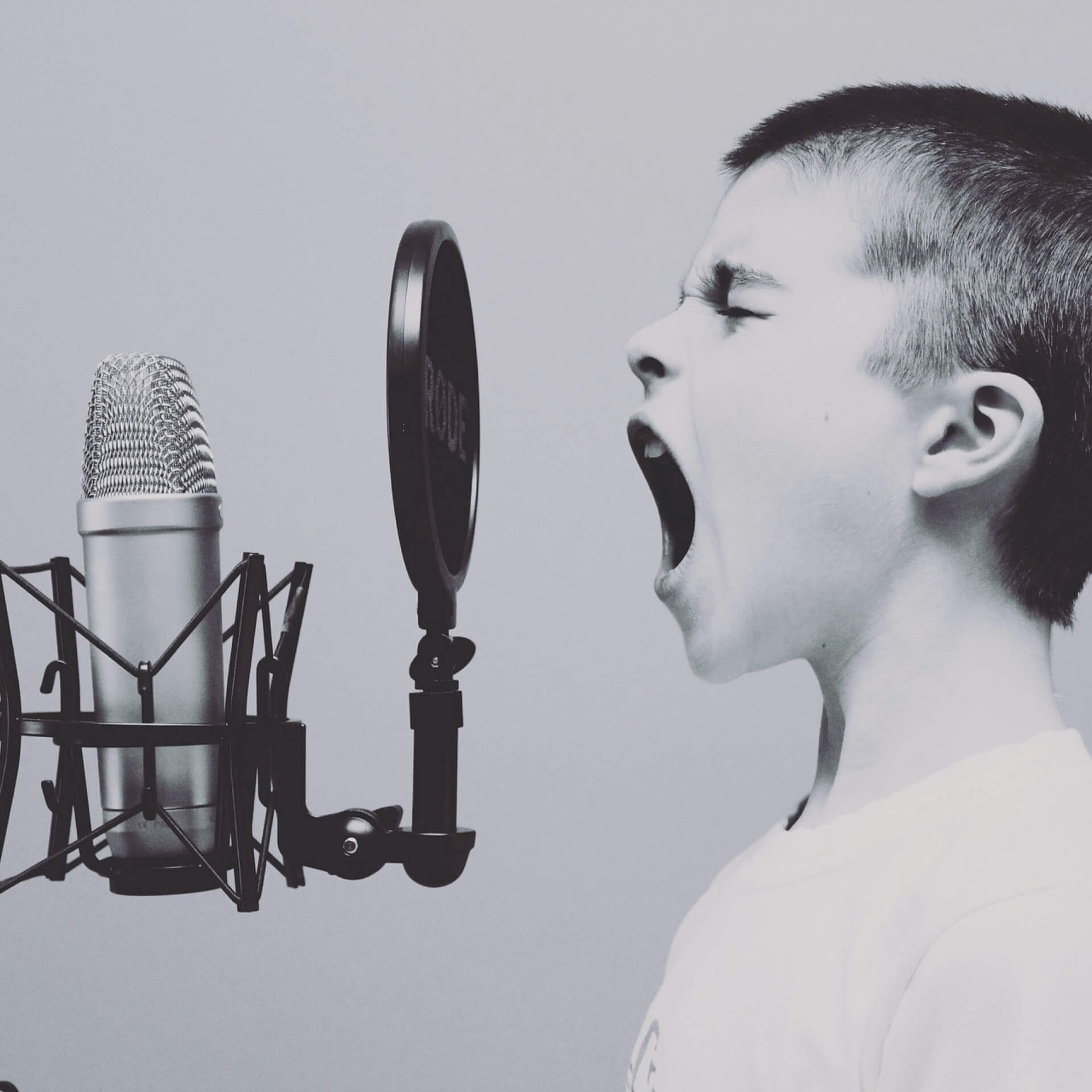 A child passionately sings or shouts into a microphone, with a pop filter in front, on a plain background.