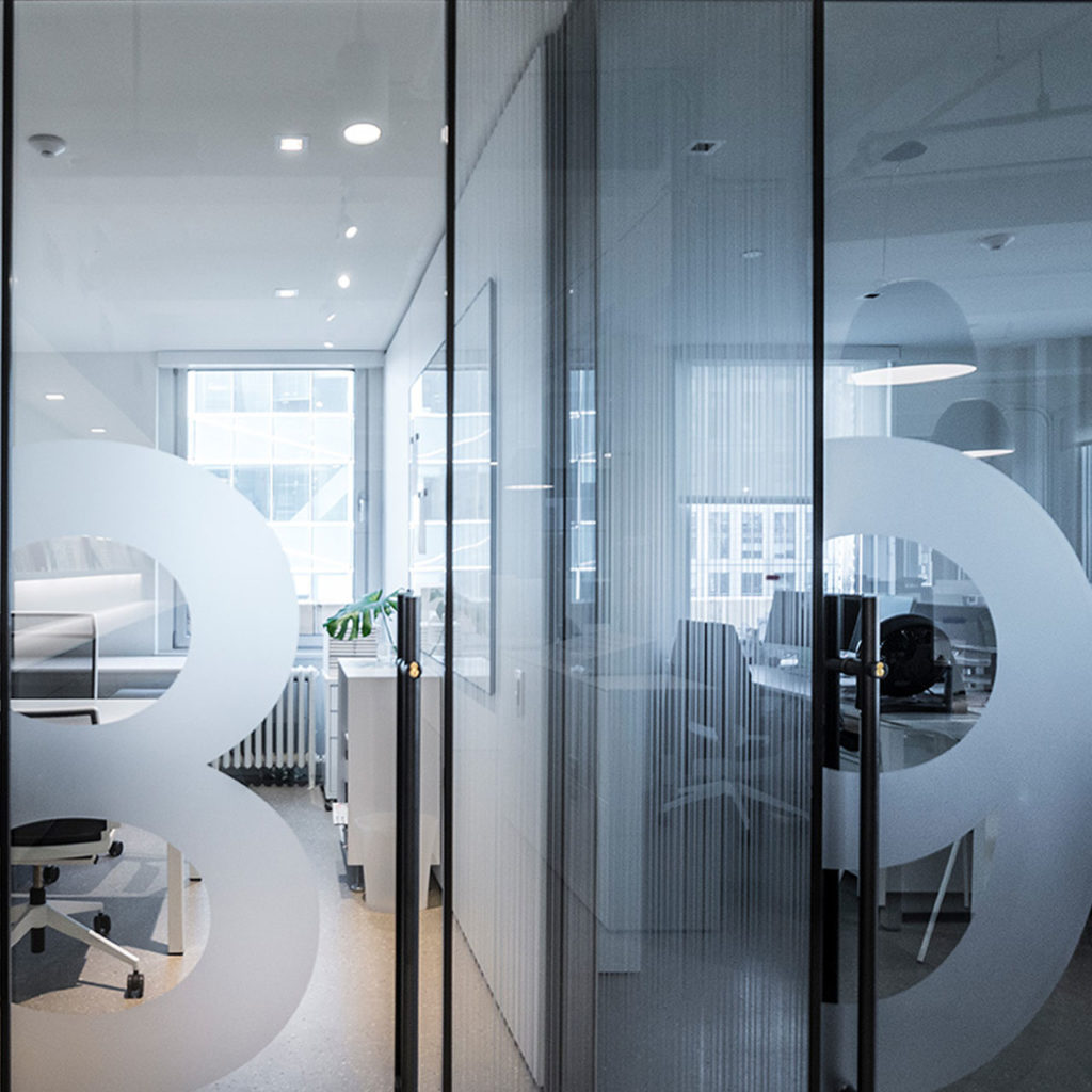 Modern office with glass walls and minimalistic design. Circular decals on glass. Inside are workstations, chairs, a small plant, and computers. Bright lighting and large windows.