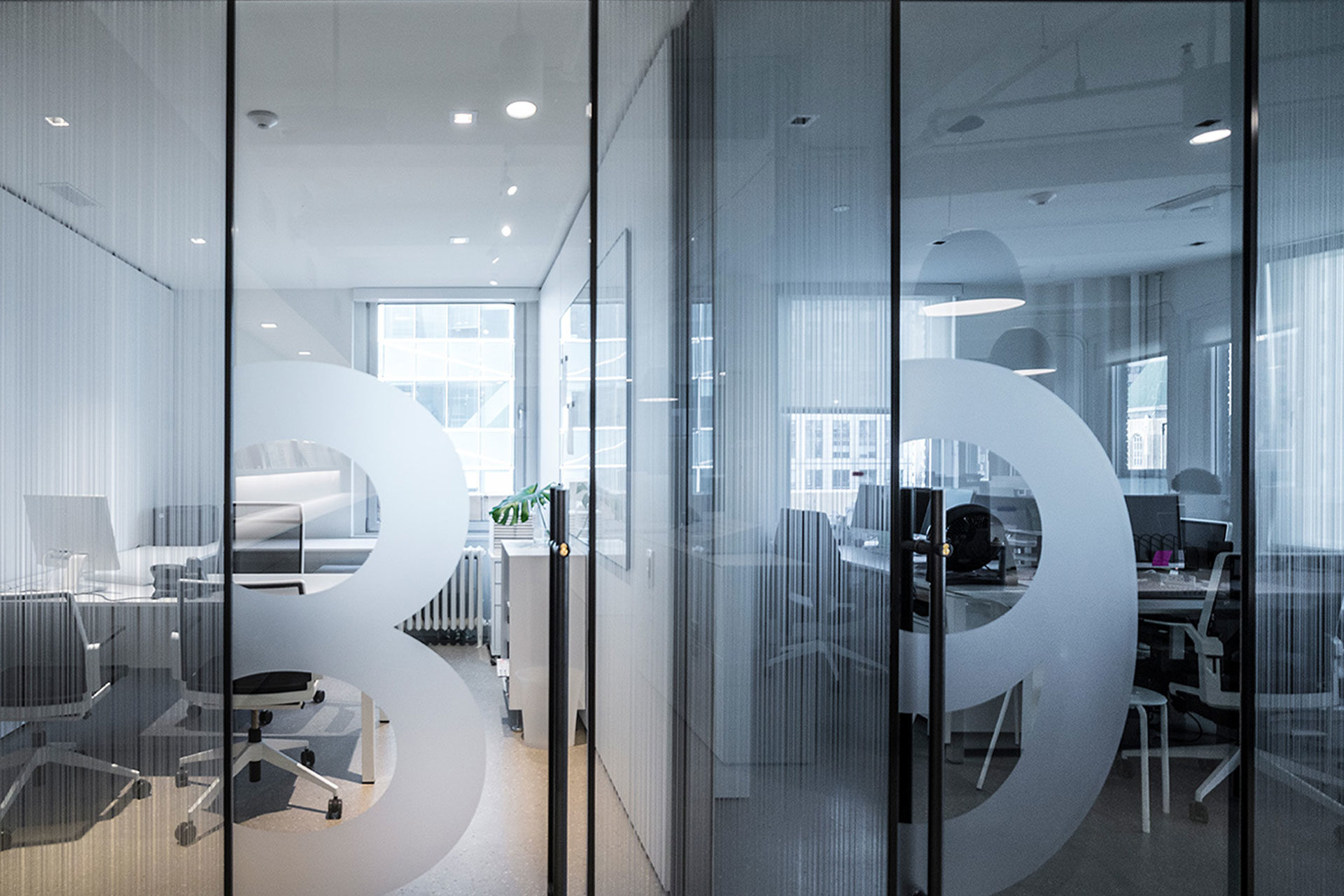 Modern office with glass walls and minimalistic design. Circular decals on glass. Inside are workstations, chairs, a small plant, and computers. Bright lighting and large windows.