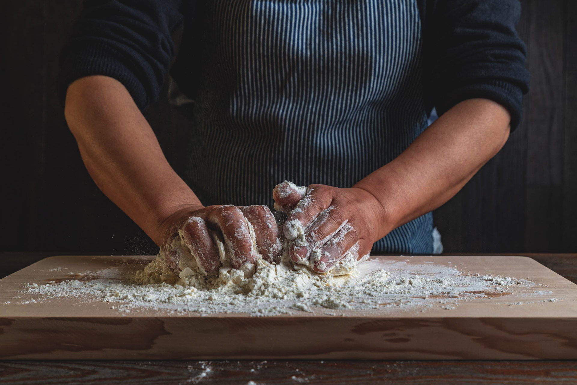 Person kneading dough with flour on a wooden board, wearing a striped apron.