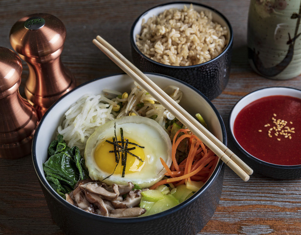 A bowl of bibimbap with vegetables and a fried egg, chopsticks on top. Sides include rice, red sauce with sesame seeds, and a ceramic container. Copper salt and pepper shakers are nearby.