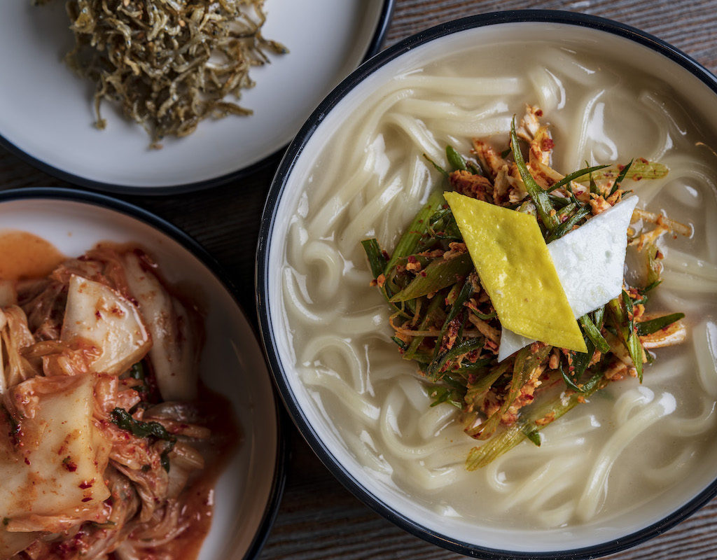 A bowl of noodle soup garnished with vegetables, a side of kimchi, and a dish of dried greens on a wooden table.