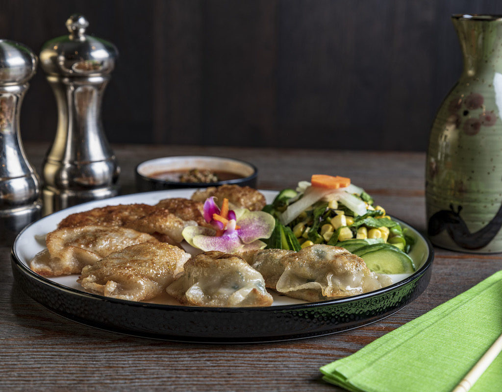 Plate of dumplings with sauce, salad, and edible flower, placed next to soy sauce dispenser, salt and pepper shakers, chopsticks, and green napkin on a wooden table.