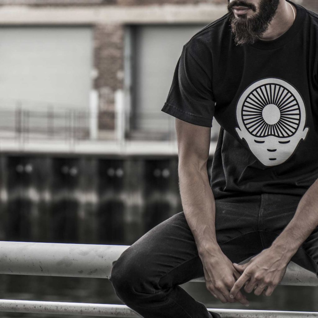 A person sits on a railing wearing a black T-shirt with a graphic design of a face and concentric circles. A waterfront and warehouses are in the background.