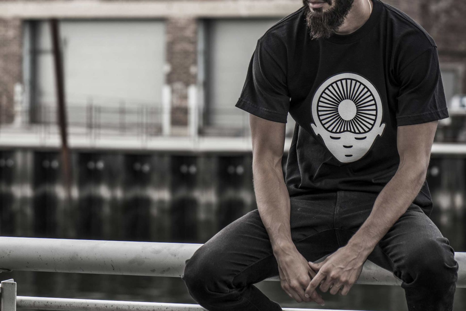 A person sits on a railing wearing a black T-shirt with a graphic design of a face and concentric circles. A waterfront and warehouses are in the background.