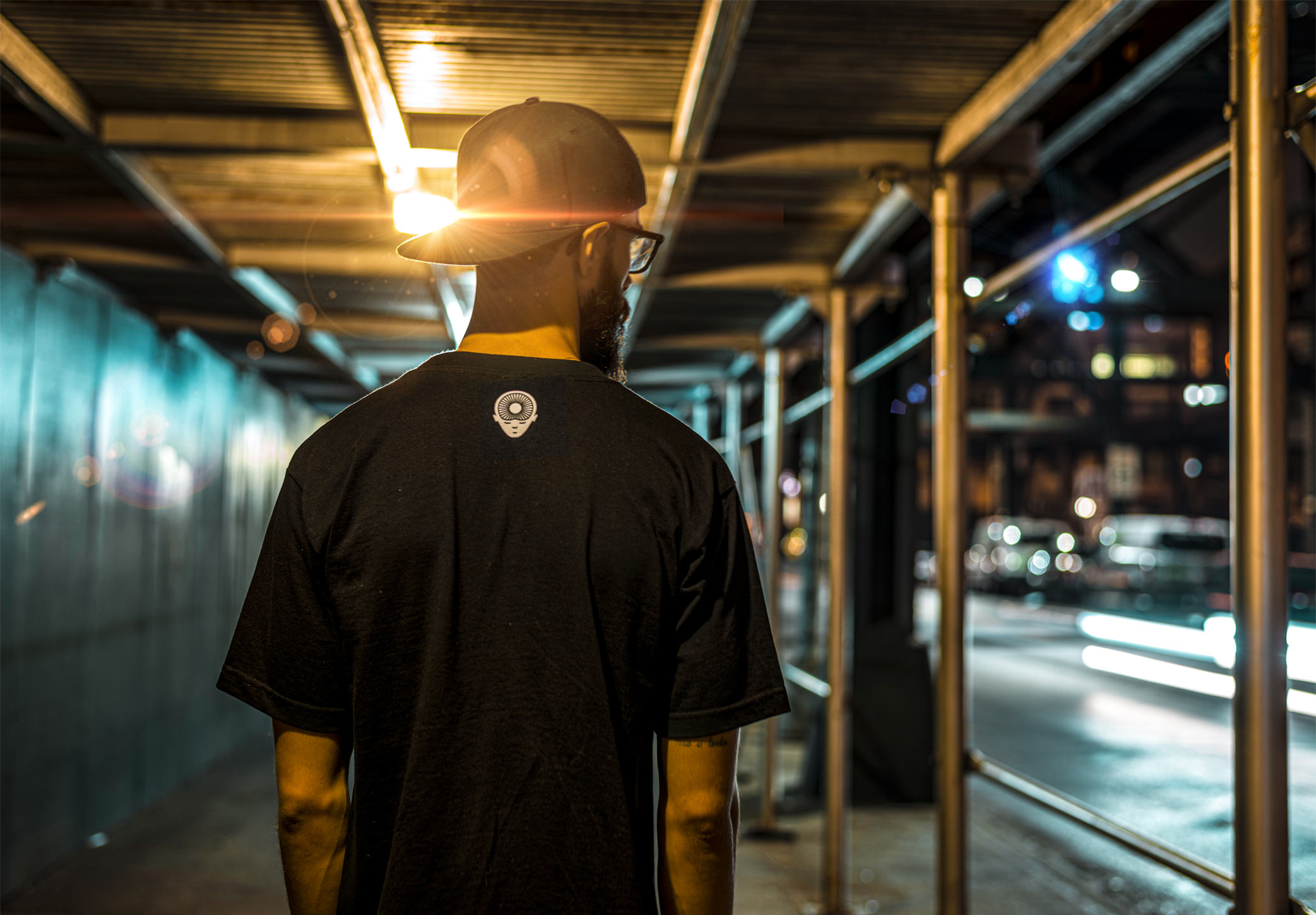 Man in a cap and a t-shirt with a light bulb design on the back, walks through a scaffolded pathway at night.