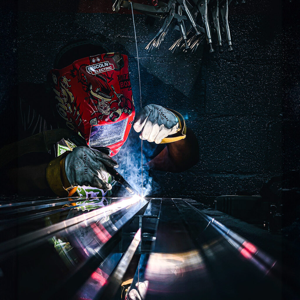 A welder wearing a red protective helmet works on metal in a dimly lit workshop, with sparks and bright light emanating from the welding process.